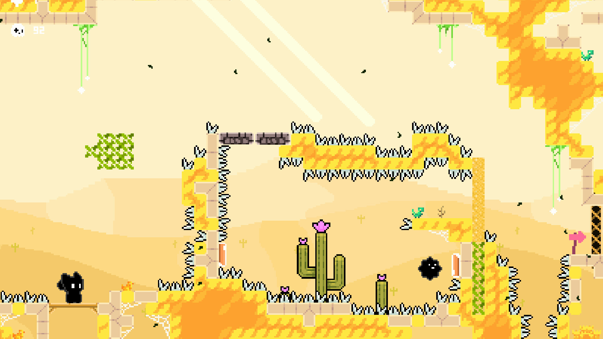 The image shows a pixel art scene from a video game, set in a desert-themed level with a yellow and beige color palette. The terrain is made of blocky platforms edged with sharp black-and-white spikes, posing obstacles for the player. In the bottom left corner, a small black character with large ears stands on a platform, facing right, while a black floating creature with a single eye hovers near cactus plants decorated with pink flowers in the middle of the scene. The background is light beige with subtle gradients, and small details like green vines, patches of grass, and scattered white sparkles add texture to the environment.
