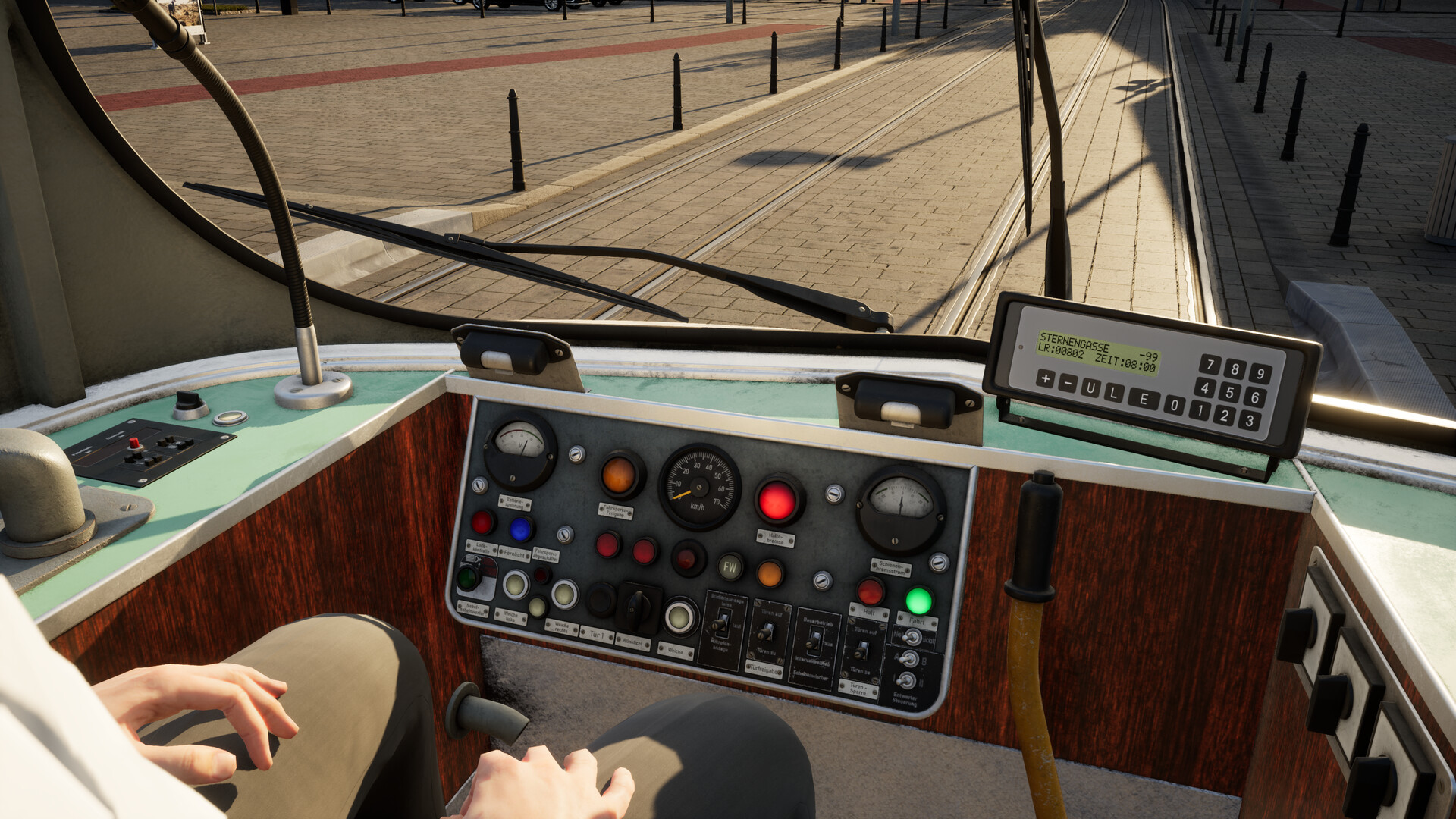 The image shows the interior of a tram driver's cabin, viewed from behind the driver, who is partially visible with hands resting on their lap. The dashboard includes numerous buttons, switches, and gauges, with a speedometer prominently displayed in the center, showing a speed of zero kilometers per hour. Outside the front window, tram tracks run straight ahead along a paved street lined with black bollards, and sunlight casts long shadows across the scene. A digital display panel on the right side of the dashboard shows text including the destination "STERNENGASSE" and timing information.
