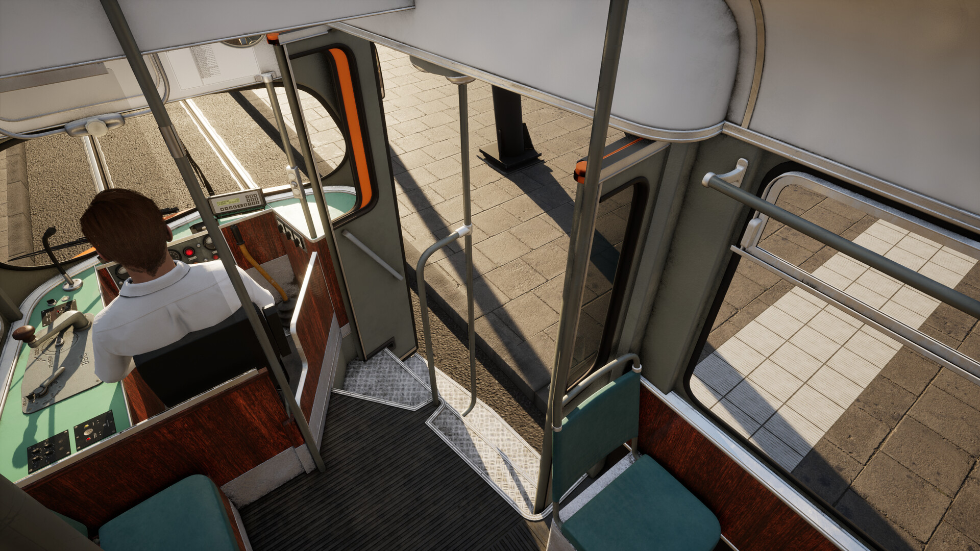 The image shows the interior of a tram or streetcar from behind the driver, who is seated at the controls wearing a white shirt. The control panel includes various levers and buttons with a green surface and wooden accents. To the right, there is an open door leading to the sidewalk with tiled pavement visible outside, along with a black street lamp casting shadows. Inside, there are teal cushioned seats and metal handrails for passengers to hold onto while standing. The scene is well-lit, suggesting daytime.