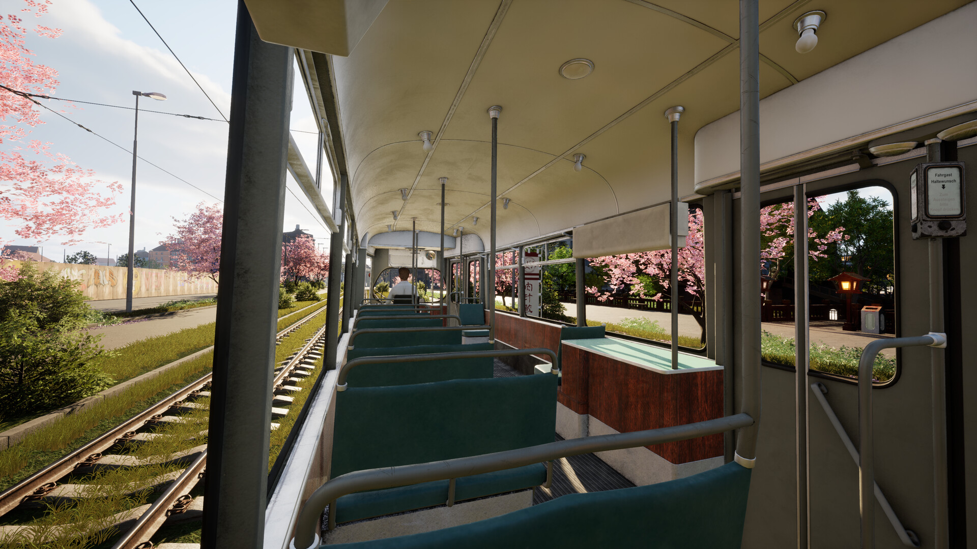 The image shows the interior of a vintage tram or streetcar with greenish-blue bench seats arranged in pairs along both sides. The ceiling is softly curved with small round lights spaced evenly along its length, and metal poles for standing passengers extend from floor to ceiling. Outside the wide windows, the tram is traveling beside grassy train tracks lined with blooming pink cherry blossom trees, while a person with short hair sits near the front of the tram facing forward. The atmosphere is bright and peaceful, with clear skies and sunlight illuminating both the interior and the scenic exterior.