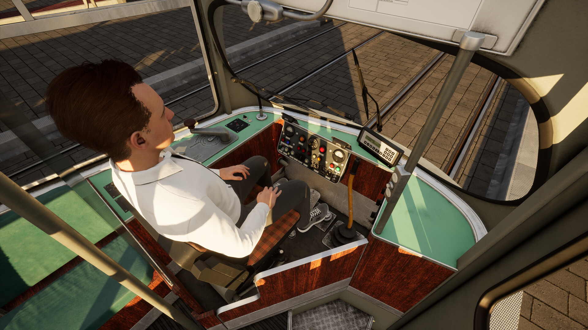 The image shows a man seated in the driver's cabin of a tram or streetcar, viewed from a high angle behind and slightly to his left. He is sitting on a patterned seat wearing a white shirt and gray pants, with his hands resting on his lap. In front of him is a control panel featuring various buttons, switches, dials, and a small digital display, along with two pedals on the floor. The tram is stationary on tracks that run parallel to a paved sidewalk visible through the front and side windows. The interior features a combination of wood paneling and light green surfaces, illuminated by daylight.