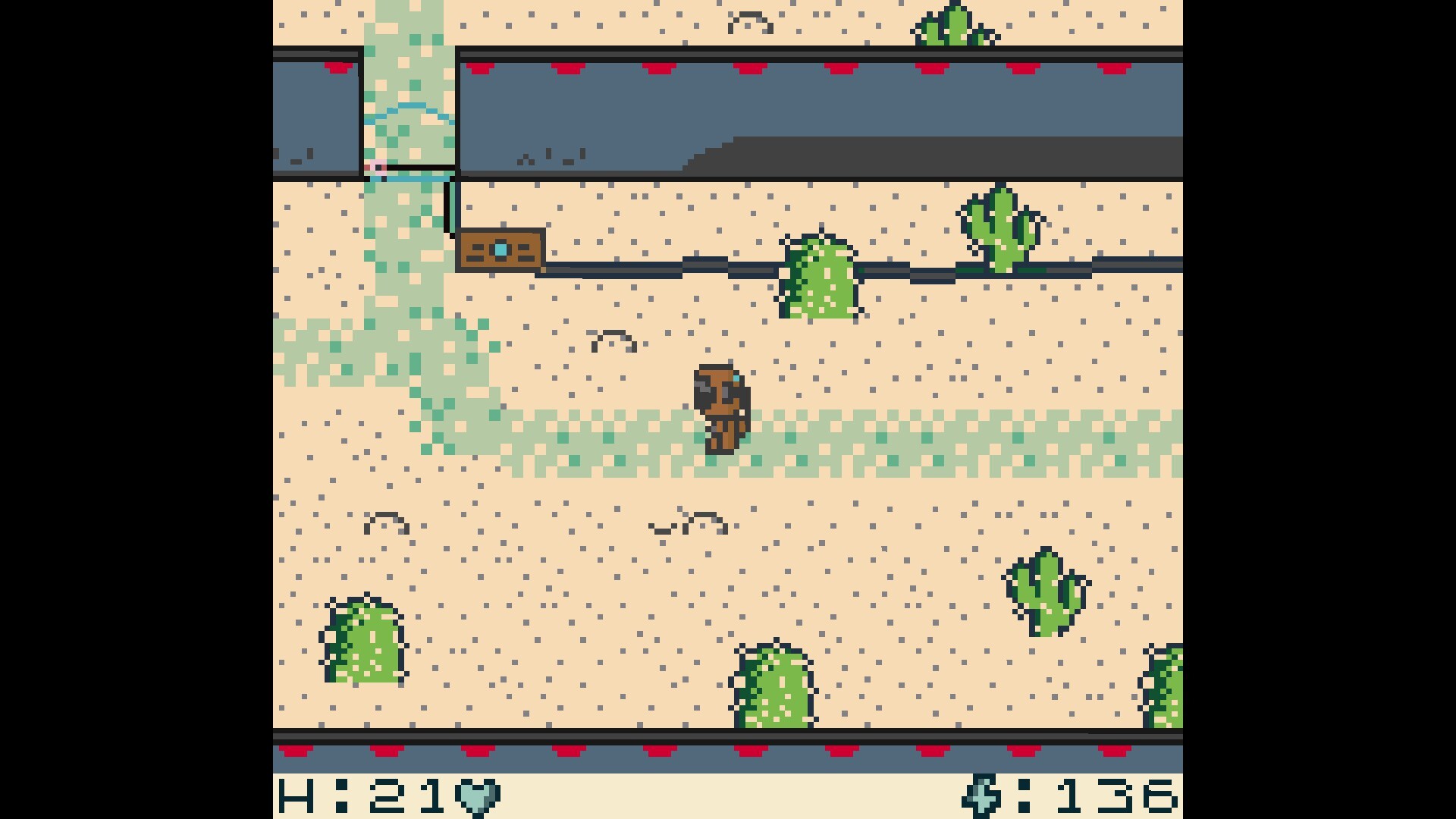 The image depicts a pixelated, top-down view of a desert-like environment from a retro-style video game. The sandy ground is dotted with green cacti and small dark specks to indicate texture. In the center, a small character dressed in brown stands on a light green path that curves upward. At the top of the screen, there is a barricade or barrier with red markings and a brown crate nearby. The bottom edge shows part of a user interface with a heart icon and numeric values, likely representing health or other stats.