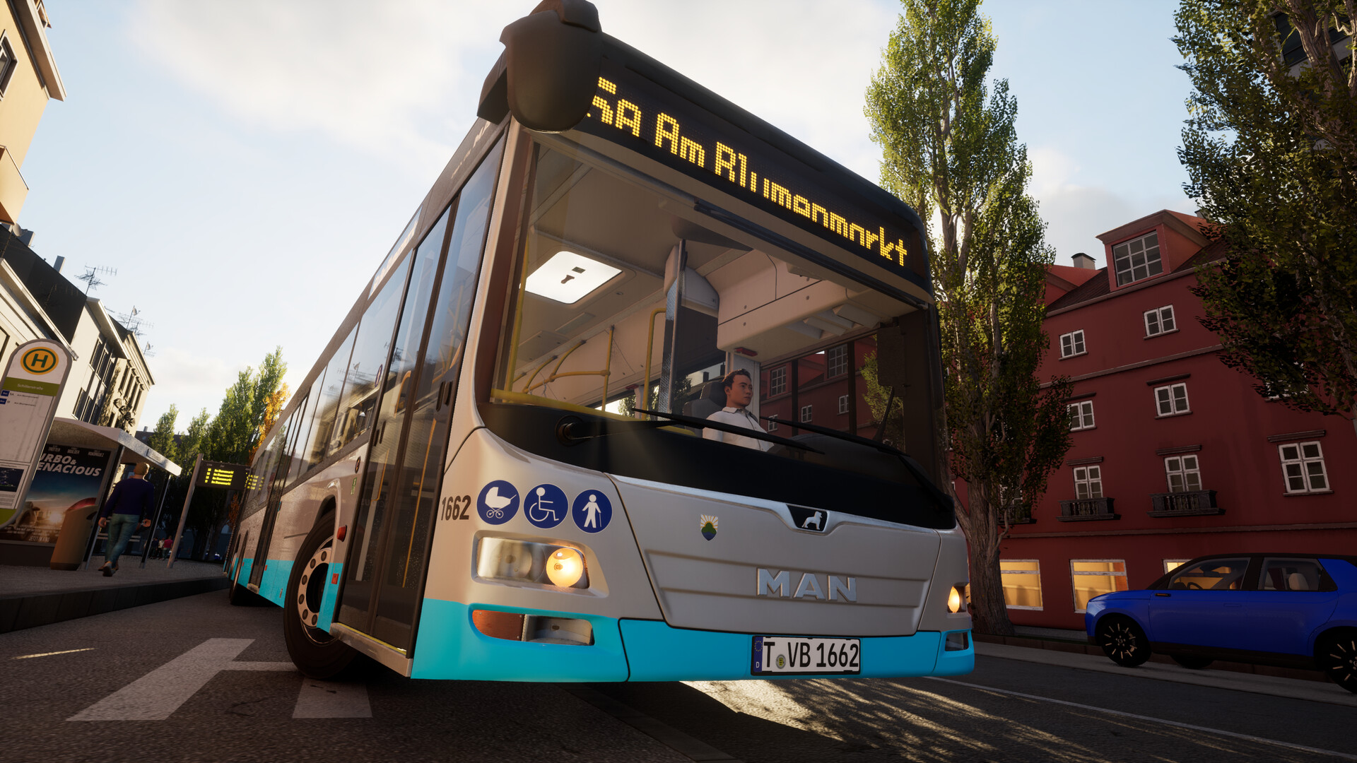 The image shows a city bus stopped at a bus stop on a quiet street lined with trees and residential buildings. The bus is white with a blue lower section and displays the route information "Sa Am Rilumammrkt" on a digital sign above the windshield. A bus driver is visible inside, sitting in the front seat, while a nearby bus stop sign and a blue car parked on the side of the road can also be seen. The scene is lit by natural daylight, suggesting it is either morning or late afternoon.