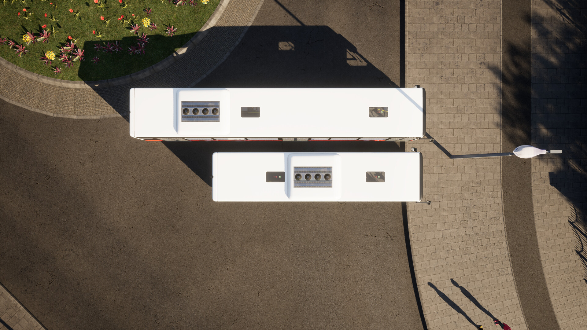 The image shows an aerial view of two white buses parked parallel to each other on a paved surface. The buses are positioned next to a curved sidewalk and a grassy area with scattered flowers and plants. Shadows cast by the buses and a nearby streetlamp extend across the pavement, and two people with long shadows are visible walking on the sidewalk below.