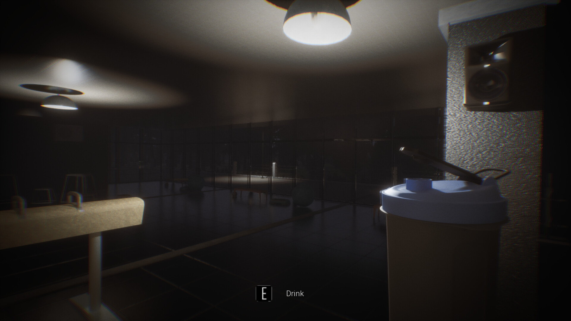 The image shows a dimly lit indoor space, likely a gym or fitness area, with tiled flooring and large glass windows along one wall. In the foreground to the right, there is a close-up of a plastic shaker bottle with a white lid and blue accents, accompanied by an on-screen prompt that reads 'E Drink.' Overhead, two ceiling lights cast soft pools of light, illuminating some gym equipment such as benches and exercise balls faintly visible in the background. The overall atmosphere is dark, with shadows dominating much of the room.